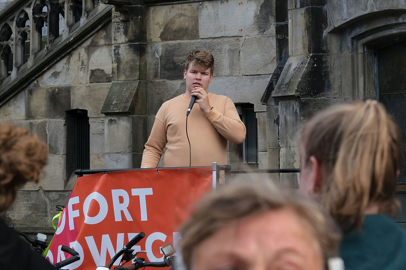 Ben Jansen spricht bei der Mahnwache vor dem Aachener Rathaus in ein Mikrofon. Vor ihm hängt ein orange-rotes Banner des Radentscheids.