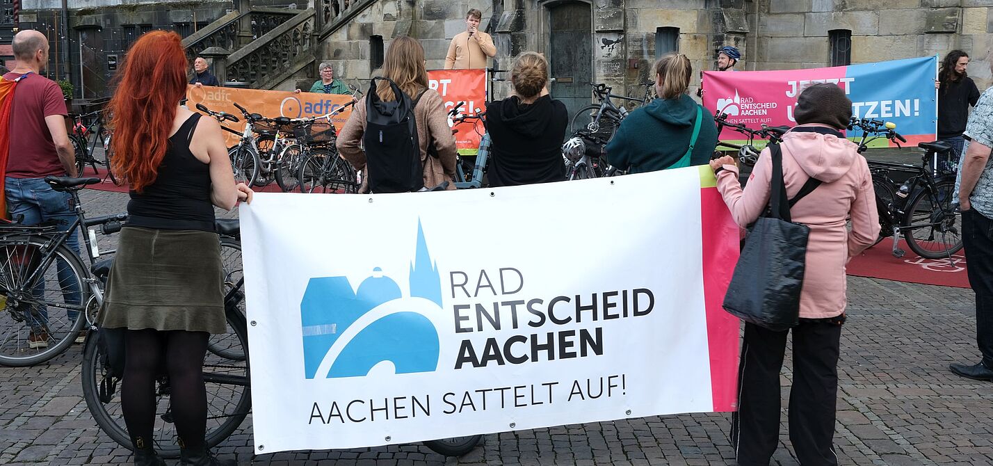 Mahnwache vor dem Rathaus Teilnehmende einer Mahnwache vor dem Aachener Rathaus stehen mit Fahrrädern und Bannern von Radentscheid Aachen und ADFC vor einem Redner mit Mikrofon.