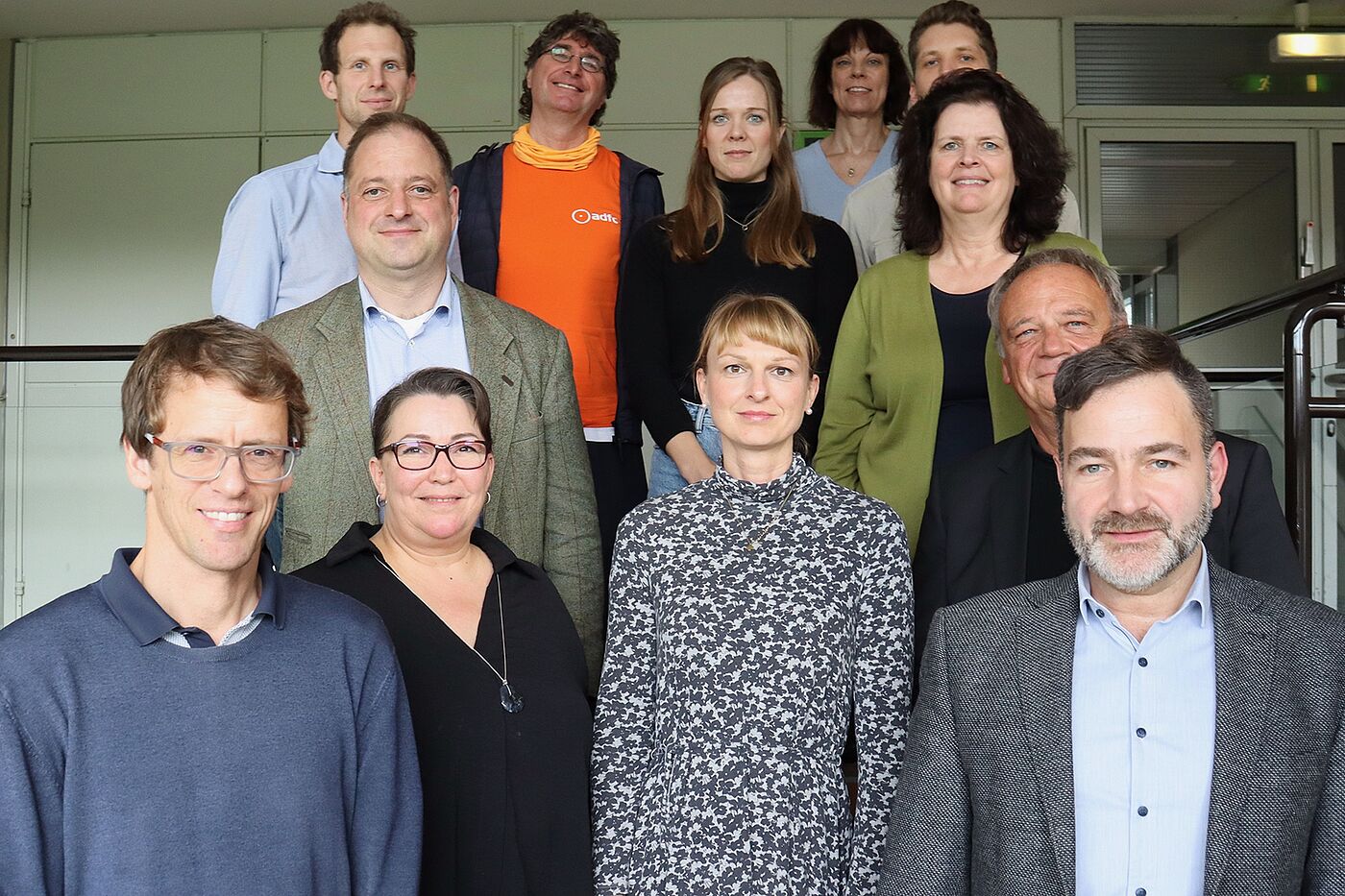 Zwölf Menschen stehen auf einem Podest. Man sieht nur ihre Oberkörper. Im Hintergrund ist eine Schrankwand zu erkennen.